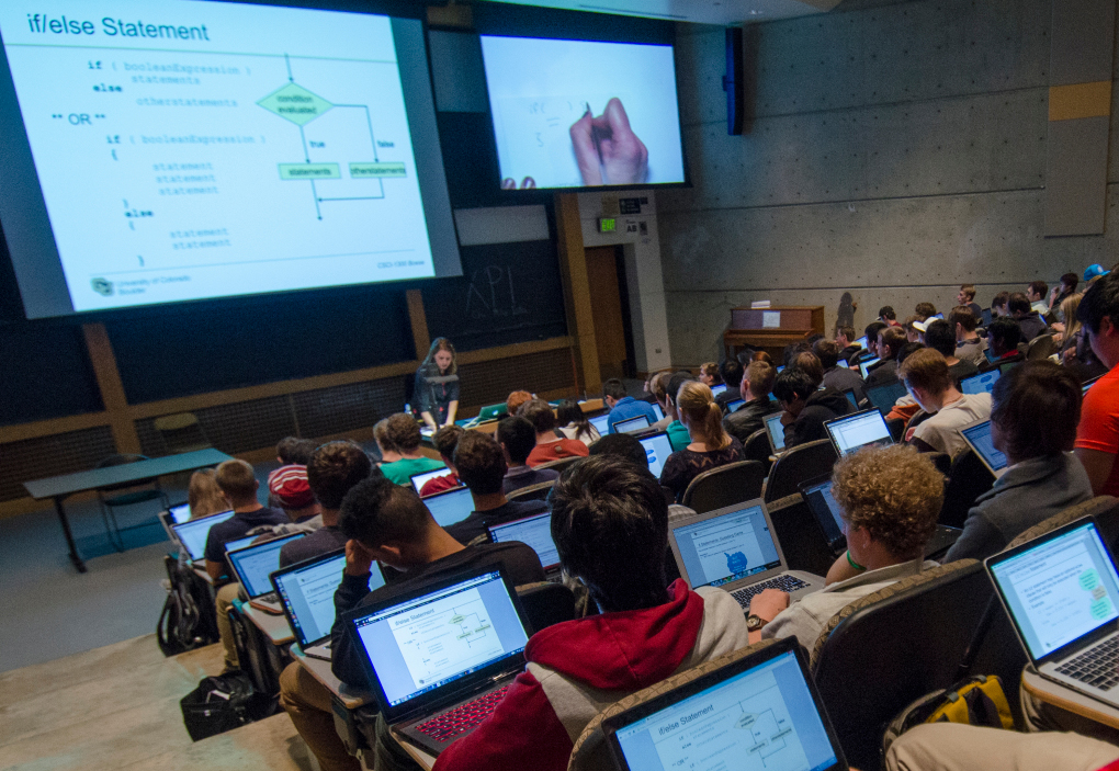 An instructor teaches using a projector in a large classroom with students who are following along on their laptops.